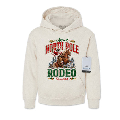 Oatmeal-colored youth pullover fleece hoodie featuring a festive North Pole Rodeo Christmas design with Cowboy Santa riding a rearing horse, snowy pine trees, vintage red and green Western lettering, December 25th details, a front kangaroo pocket, and ribbed cuffs and hem.