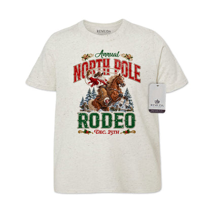 Oatmeal-colored youth short sleeve T-shirt featuring a festive North Pole Rodeo Christmas design with Cowboy Santa riding a rearing horse, snowy pine trees, vintage red and green Western lettering, December 25th details, and a classic crewneck style.