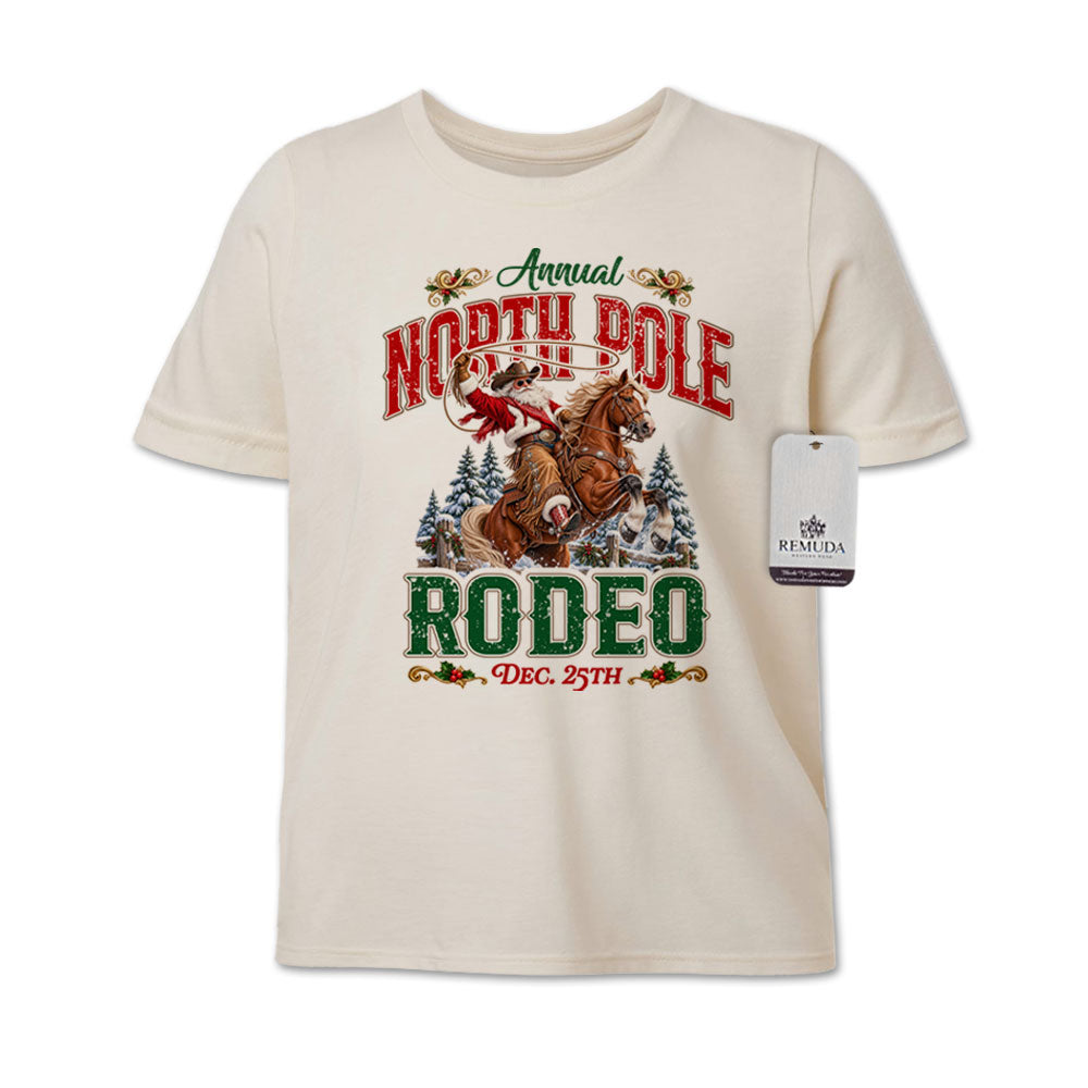 Solid natural-colored youth short sleeve T-shirt featuring a festive North Pole Rodeo Christmas design with Cowboy Santa riding a rearing horse, snowy pine trees, vintage red and green Western lettering, December 25th details, and a classic crewneck style.