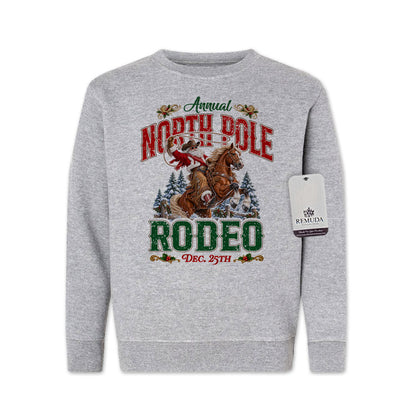 Heather gray youth crewneck fleece sweatshirt featuring a festive North Pole Rodeo Christmas design with Cowboy Santa riding a rearing horse, snowy pine trees, vintage red and green Western lettering, December 25th details, and ribbed cuffs and hem.